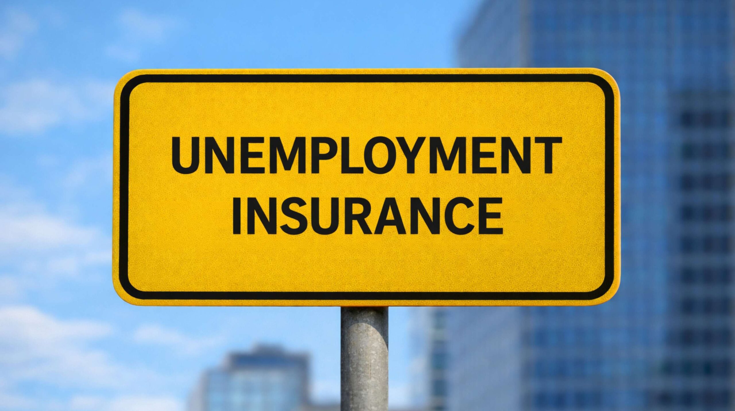 Yellow road-style sign with the words “UNEMPLOYMENT INSURANCE” in bold black letters, mounted on a metal pole, with blurred office buildings in the background.