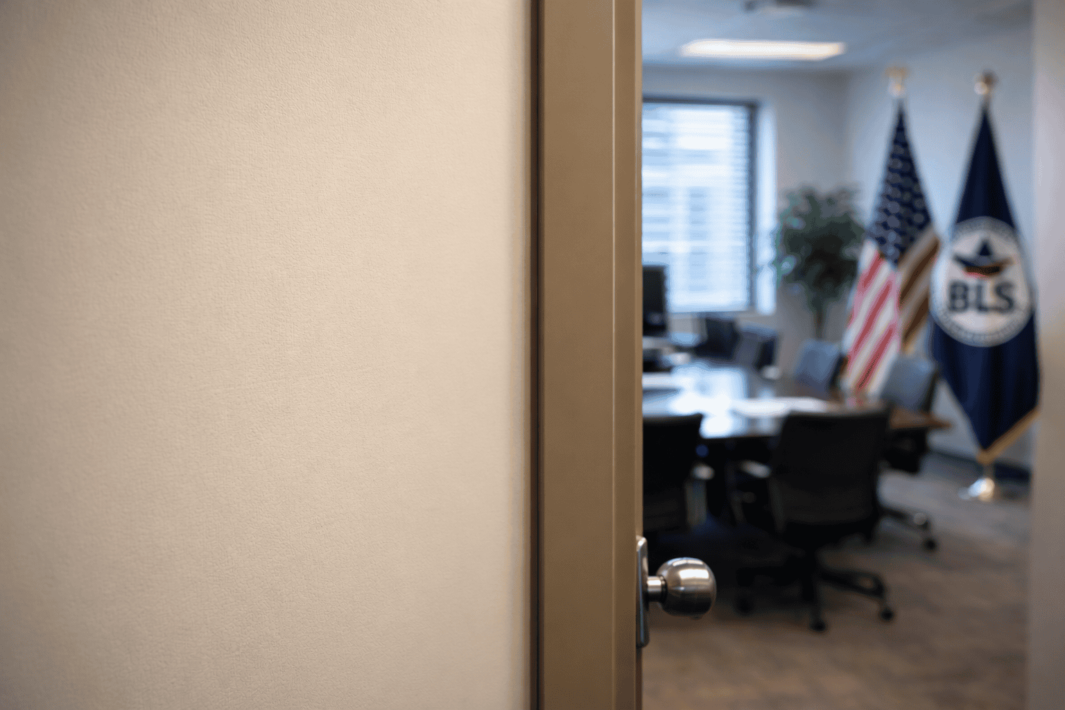 Porta de escritório parcialmente aberta mostrando uma sala de reunião desfocada ao fundo, com mesa, cadeiras e bandeiras, iluminada por luz natural entrando pela janela.