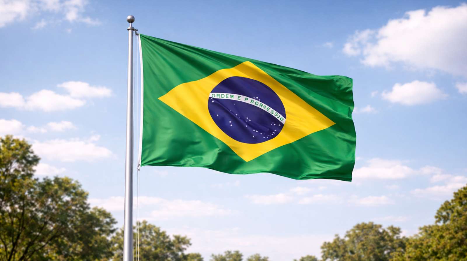 Bandeira do Brasil tremulando em um mastro sob céu azul com nuvens, com o símbolo nacional em verde, amarelo e azul.