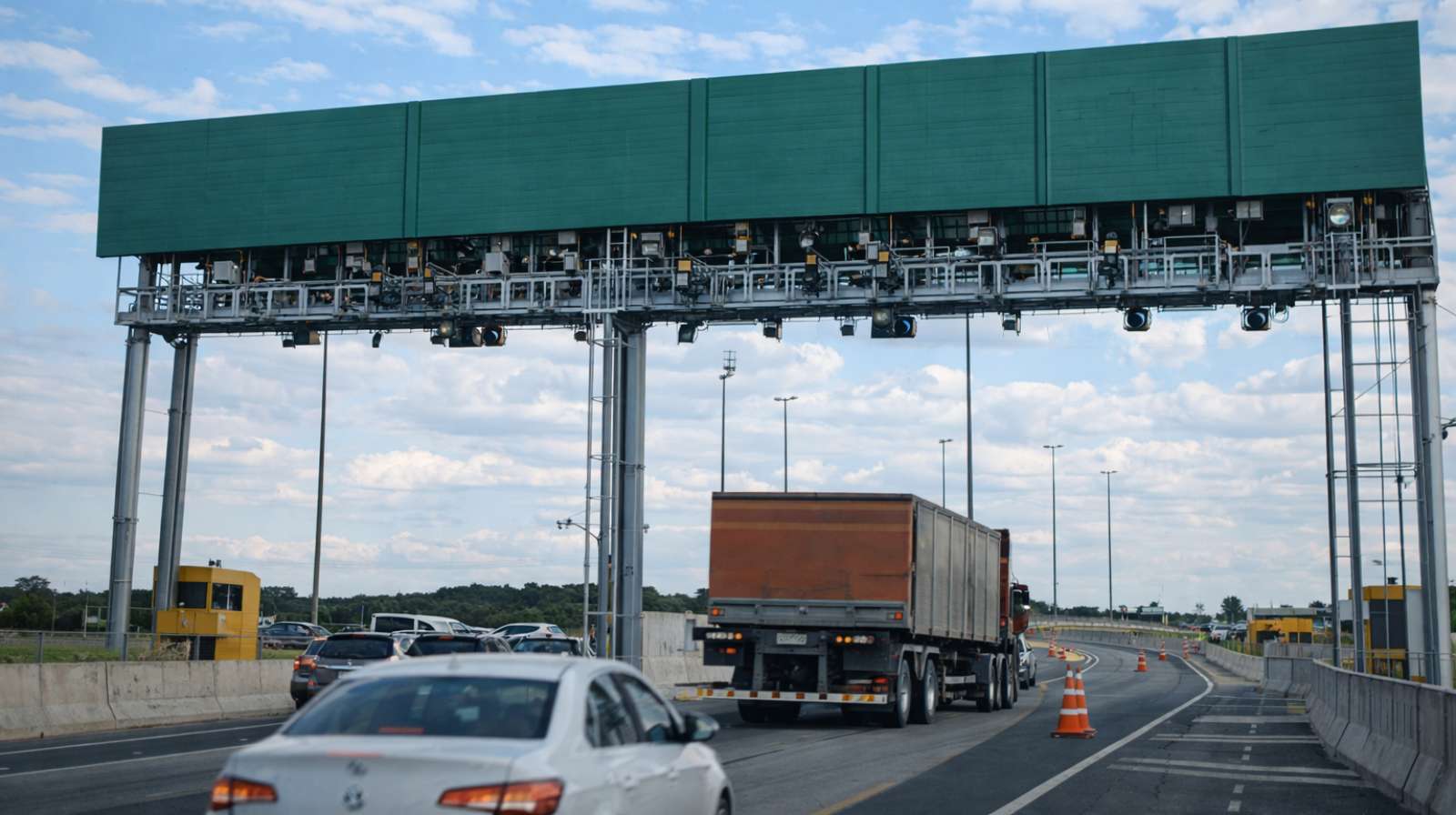 Pórtico de pedágio eletrônico sobre rodovia com carros e caminhão em movimento, cones de sinalização e céu parcialmente nublado ao fundo.