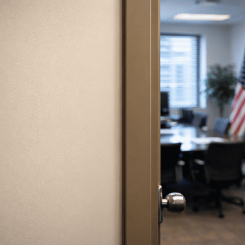 Porta de escritório parcialmente aberta mostrando uma sala de reunião desfocada ao fundo, com mesa, cadeiras e bandeiras, iluminada por luz natural entrando pela janela.