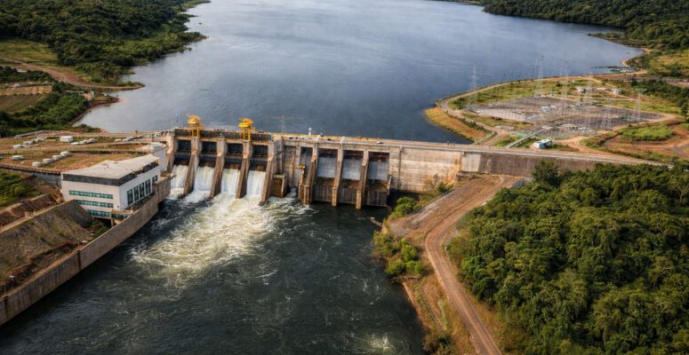 Vista aérea da Usina Hidrelétrica de Colíder, com barragem, vertedouros em operação, reservatório ao fundo e área de vegetação ao redor.