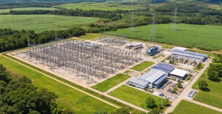 Subestação de energia elétrica vista de cima, cercada por áreas verdes e campos agrícolas, com torres de transmissão e linhas de alta tensão em paisagem rural sob céu parcialmente nublado.
