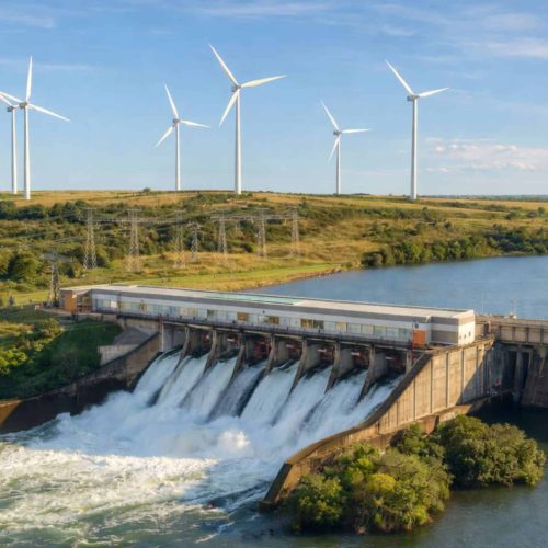 Usina hidrelétrica com vertedouro em operação e água sendo liberada, com torres de transmissão e aerogeradores ao fundo em paisagem rural sob céu azul.