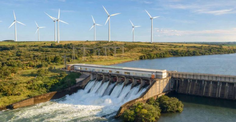Usina hidrelétrica com vertedouro em operação e água sendo liberada, com torres de transmissão e aerogeradores ao fundo em paisagem rural sob céu azul.