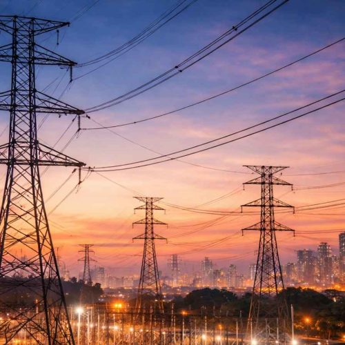 Linhas de transmissão de energia elétrica e torres de alta tensão próximas a uma subestação com skyline urbano ao fundo durante o pôr do sol.