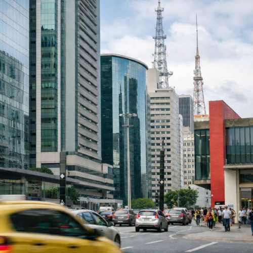 Rua movimentada em centro financeiro brasileiro com prédios comerciais, carros no trânsito e pessoas caminhando nas calçadas.