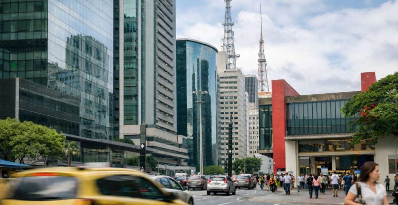 Rua movimentada em centro financeiro brasileiro com prédios comerciais, carros no trânsito e pessoas caminhando nas calçadas.