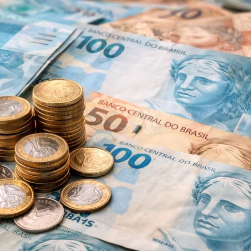 Notas e moedas de real brasileiro espalhadas sobre uma mesa, com destaque para cédulas de 100 e 50 reais e moedas empilhadas.