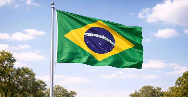 Bandeira do Brasil tremulando em um mastro sob céu azul com nuvens, com o símbolo nacional em verde, amarelo e azul.