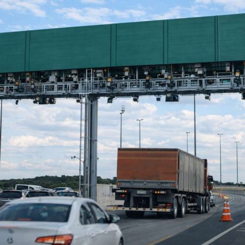 Pórtico de pedágio eletrônico sobre rodovia com carros e caminhão em movimento, cones de sinalização e céu parcialmente nublado ao fundo.