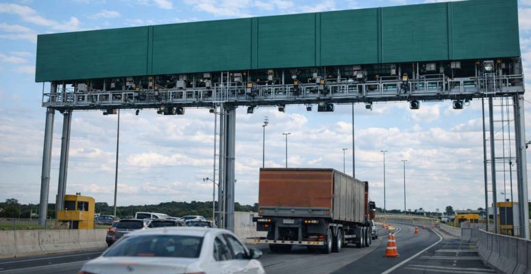 Pórtico de pedágio eletrônico sobre rodovia com carros e caminhão em movimento, cones de sinalização e céu parcialmente nublado ao fundo.