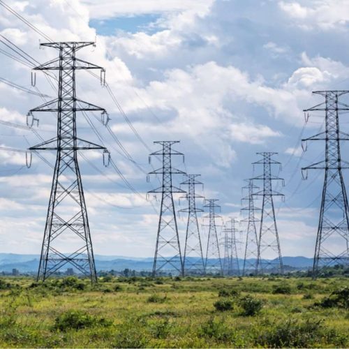 Linhas de transmissão de energia em longa distância atravessando área rural sob céu parcialmente nublado, representando infraestrutura do setor elétrico.