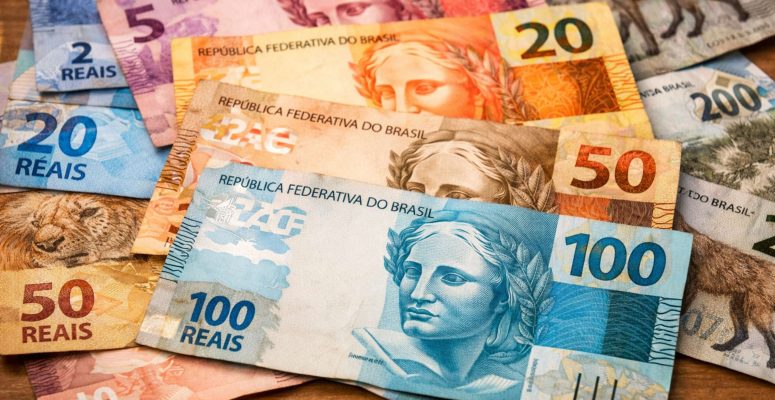 Notas do real brasileiro de 2, 5, 10, 20, 50, 100 e 200 reais espalhadas sobre uma mesa de madeira, com destaque para a cédula azul de 100 reais em primeiro plano.