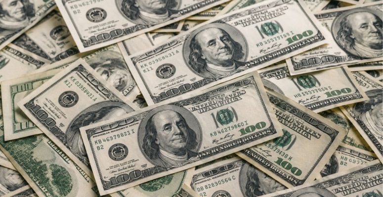 Montanha de notas de 100 dólares espalhadas e sobrepostas, destacando o rosto de Benjamin Franklin em várias cédulas, em uma composição que remete ao mercado financeiro, câmbio e fluxo internacional de capital.