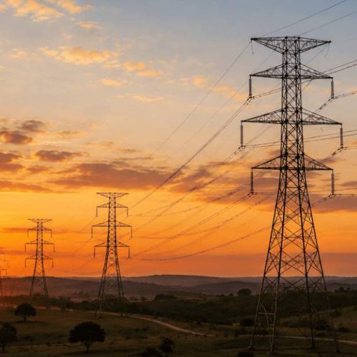 Rede de transmissão de energia elétrica com torres e linhas de alta tensão ao pôr do sol em área rural no Brasil.